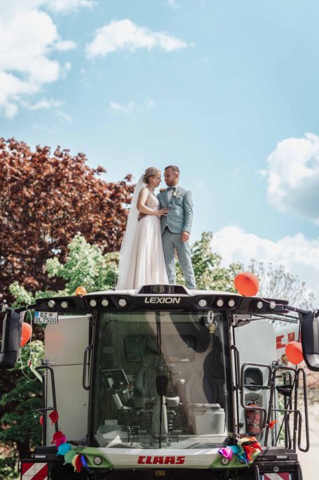 Brautpaar steht auf einem Traktor unter blauem Himmel mit Wolken und bunten Ballons.