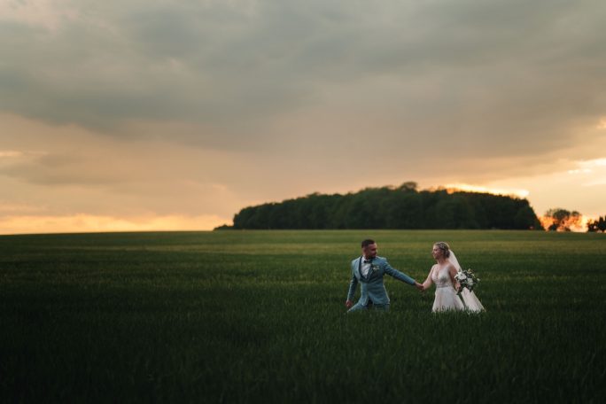 Ein Brautpaar hält sich in einem weiten grünen Feld bei Sonnenuntergang an den Händen.