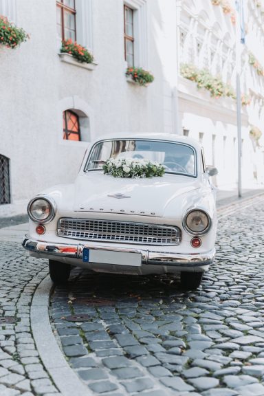 Weißes Oldtimer-Auto steht auf einer gepflasterten Straße, mit Blumen dekoriert.
