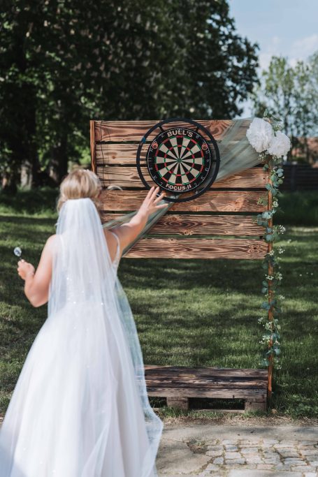 Braut wirft Dartpfeil auf eine Holzscheibe in einer grünen Umgebung.