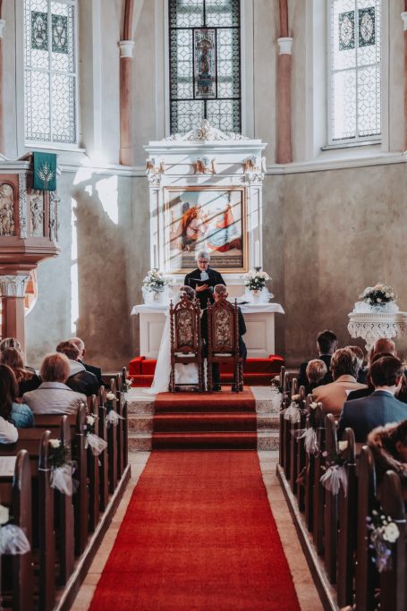 Brautpaar steht vor dem Altar in einer Kirche, Gäste sitzen auf Bänken.