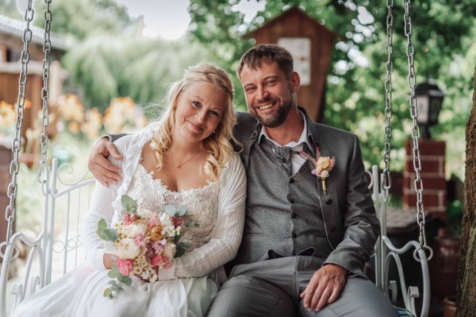 Hochzeitsfoto eines Paares auf einer Schaukel, umgeben von Natur, mit Blumen im Brautstrauß.