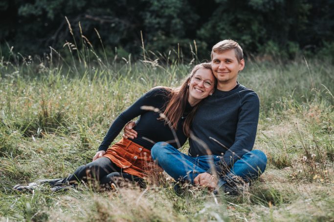 Paar sitzt im hohen Gras, lächelnd und entspannt, umgeben von Natur.