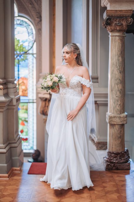 Braut in einem eleganten weißen Kleid mit Blumenstrauß, steht vor einer Fensterlandschaft.