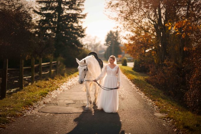 Braut in weißem Kleid führt ein weißes Pferd auf einem Weg im Herbst.