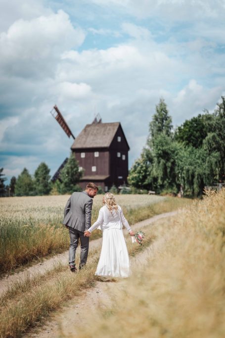 Brautpaar, Hand in Hand, auf einem Feldweg mit einer alten Windmühle im Hintergrund.