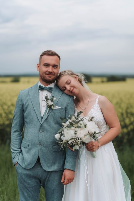 Ein Brautpaar steht lächelnd im Freien, umgeben von blühenden Feldern.