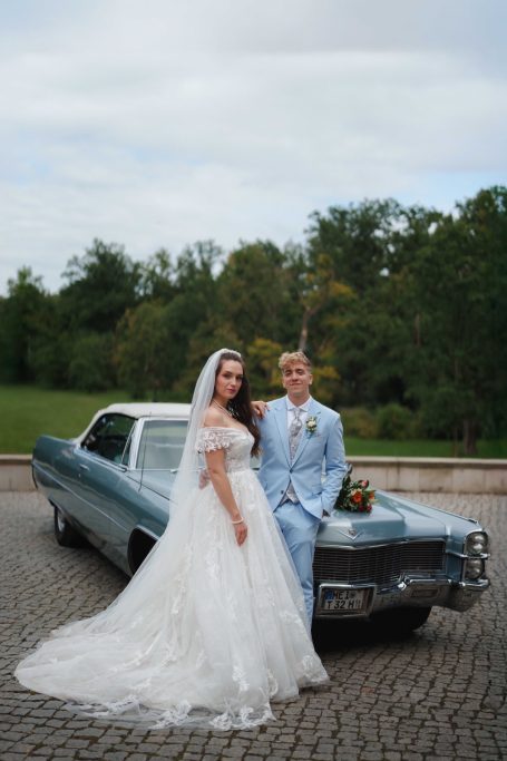Braut und Bräutigam in Hochzeitskleidung stehen neben einem klassischen Auto im Freien.