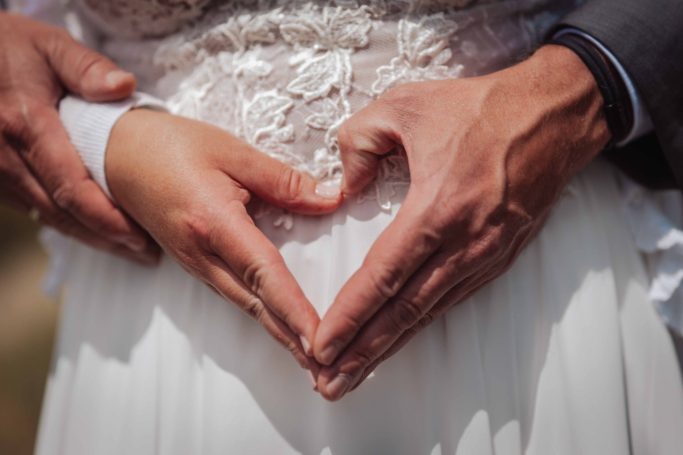 Hände bilden ein Herz auf dem Bauch einer schwangeren Frau in einem Brautkleid.