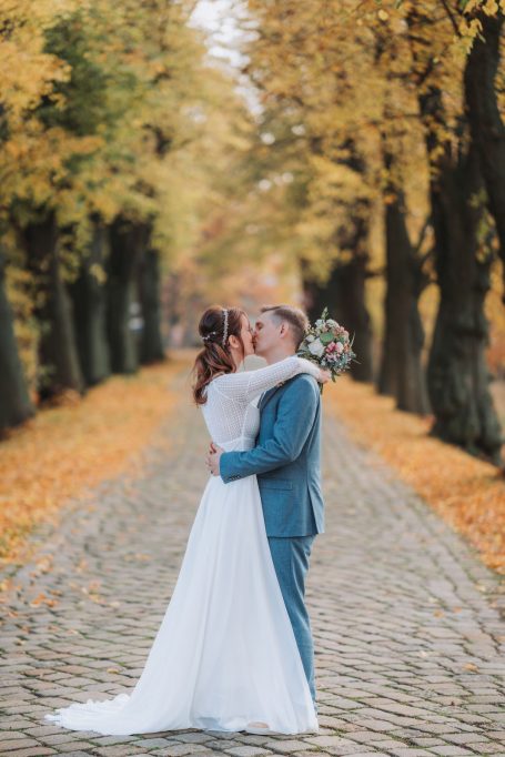 Ein Brautpaar umarmt sich auf einem gepflasterten Weg, umgeben von herbstlichen Bäumen.