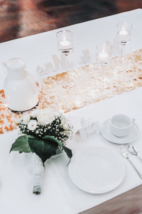 Tischdeko mit weißem Geschirr, Blumenarrangement und Kerzen auf weißem Tisch.