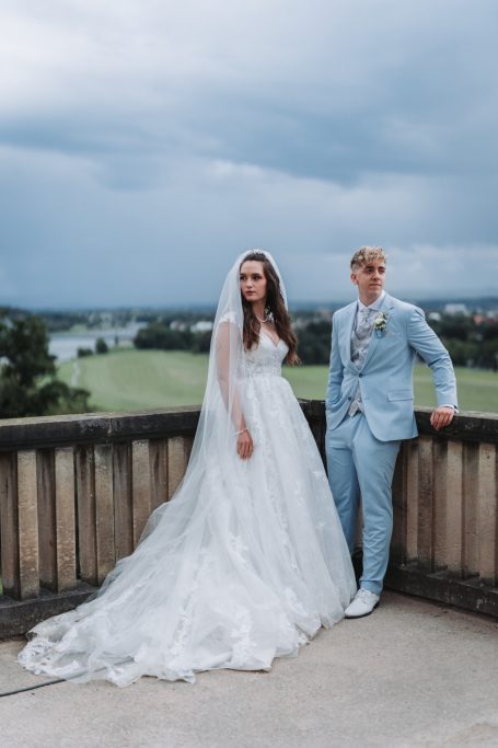 Braut in weißem Kleid und Bräutigam im hellblauen Anzug stehen vor einem grünen Landschaftshorizont.