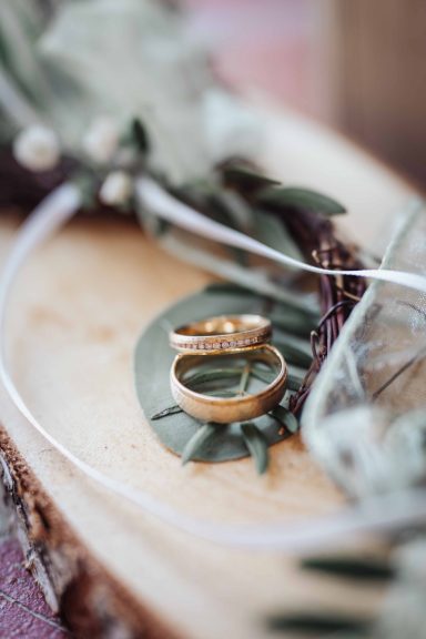 Zwei goldene Ringe auf einem Blatt und Holzuntergrund, umgeben von Pflanzen.