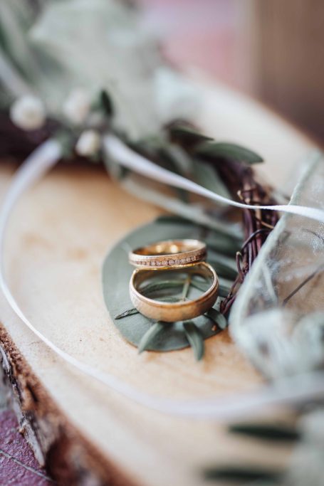 Zwei goldene Ringe auf einem Blatt und Holzuntergrund, umgeben von Pflanzen.