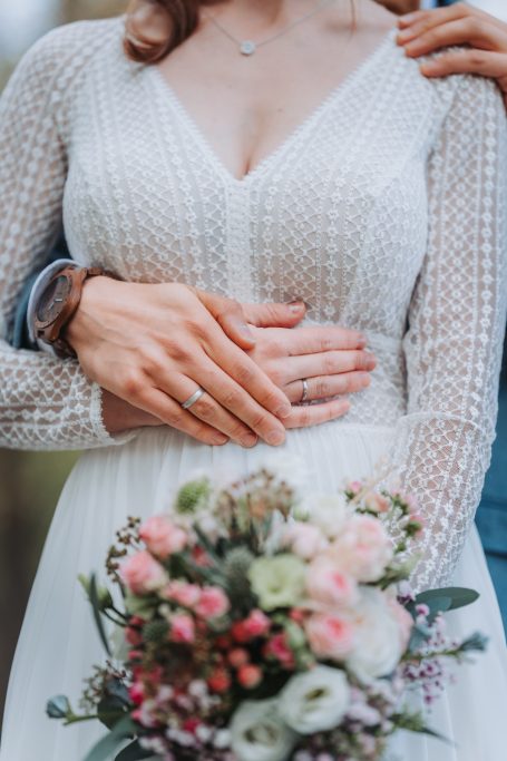 Braut in weißem Kleid mit Blumenstrauß, umarmt von einem Partner.