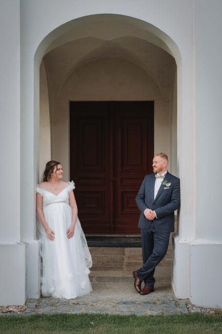 Braut in weißem Kleid und Bräutigam in Anzug stehen entspannt vor einer Holztür.