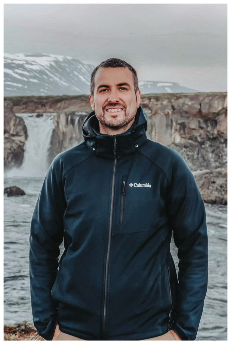 Mann mit schwarzer Jacke steht vor einem Wasserfall und Bergen. Schwarz-Weiß-Foto.