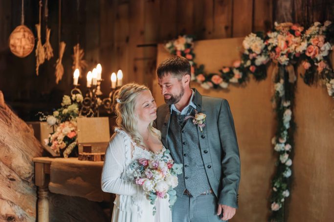 Braut und Bräutigam stehen lächelnd vor festlich dekoriertem Hintergrund.