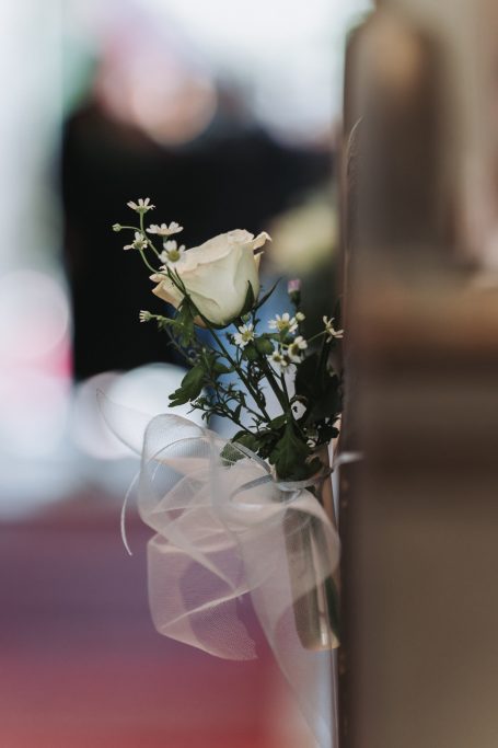 Weiße Rose mit kleinen Blumen und Schleife, dekorativ an einer Bank angebracht.