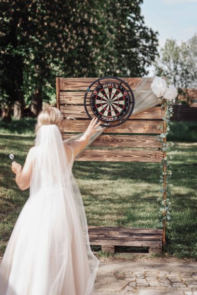 Eine Braut wirft Pfeile auf eine Dartscheibe vor einer Holzwand im Freien.
