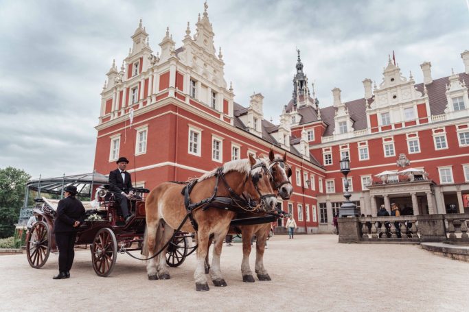 Ein Kutschenfahrzeug mit zwei Pferden vor einem prachtvollen roten Schloss.