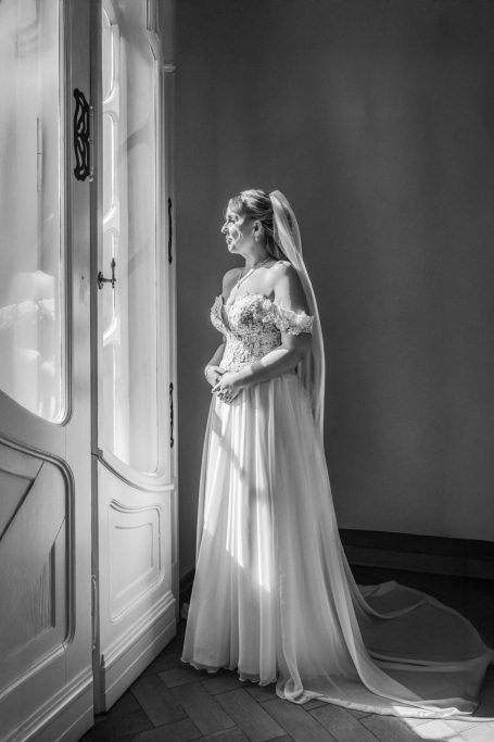 Braut in einem eleganten Hochzeitskleid steht am Fenster und schaut nach draußen.